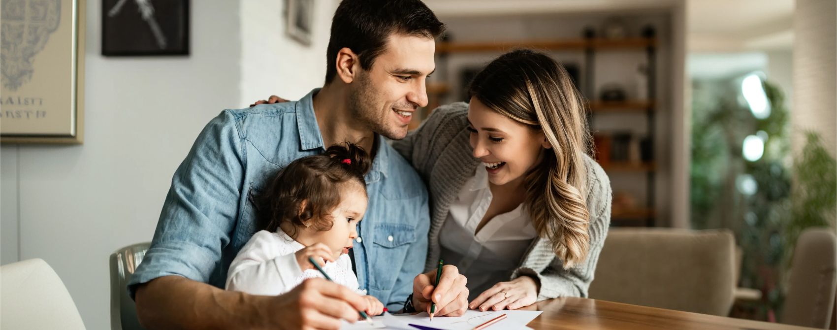 Homem e mulher sorriem enquanto acompanham uma criança pequena desenhando em folhas de papel sobre uma mesa de madeira. O homem usa camisa jeans, a mulher veste camisa branca com cardigã cinza, e o ambiente é uma sala de estar integrada à cozinha.